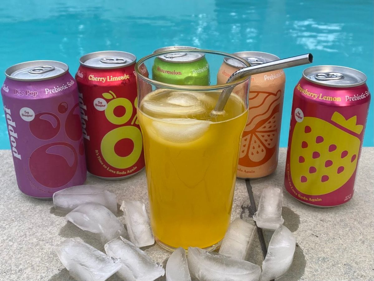 A glass of Poppi’s Orange soda surrounded by their Doc Pop, Cherry Limeade, Watermelon, Orange Crème and Strawberry Lemon cans.